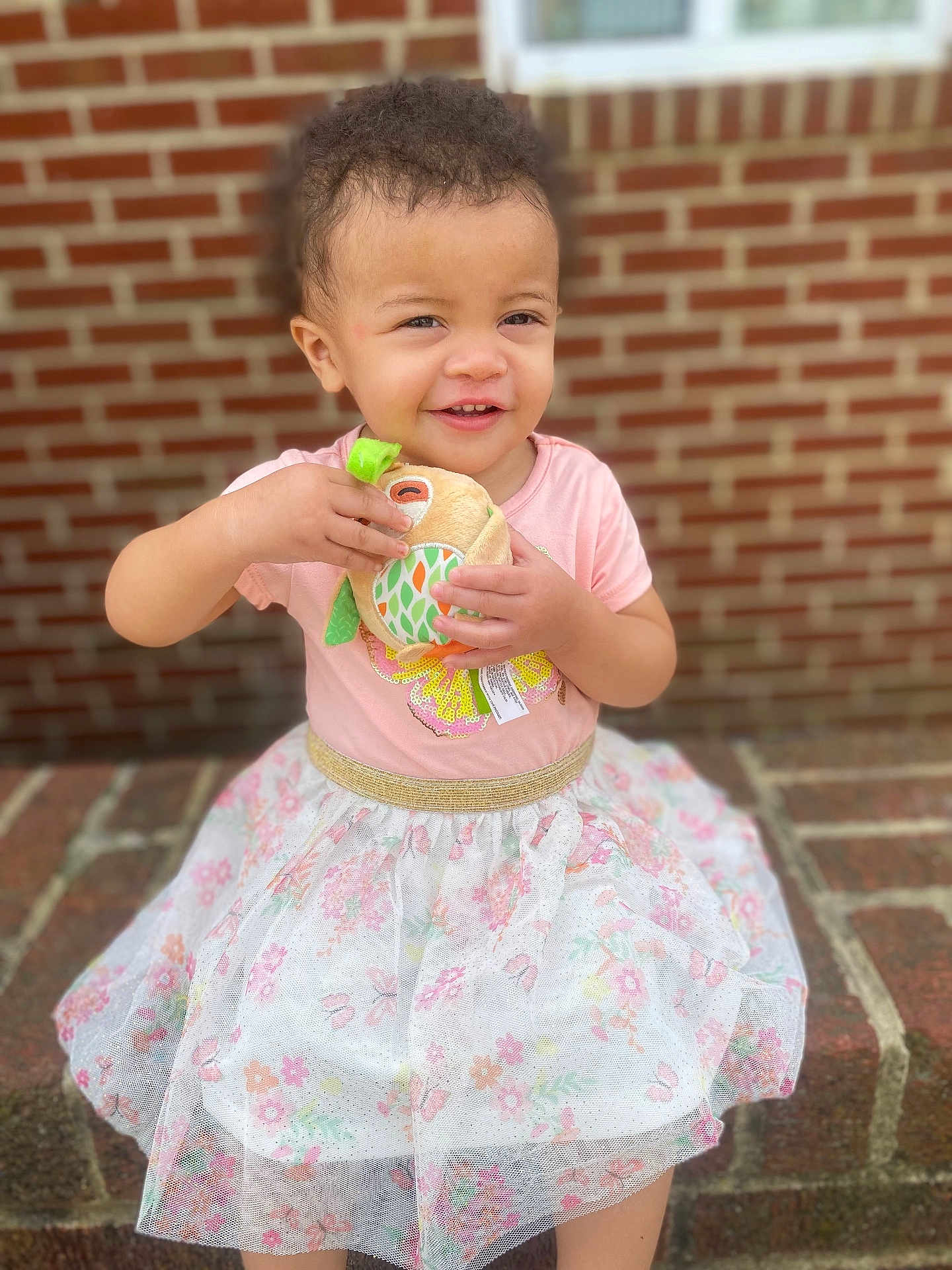 Zamaya is registered to the contest to win money with this photo: toddler, child, smiling, pink_shirt, floral_skirt, plush_toy, brick_wall, outdoor, happy, cute, sitting, daylight, person, toy, playful, young_child, casual_clothing, portrait, baby, innocence