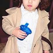 Naïla a rejoint le concours — aidez-le/la à gagner de superbes lots ! beige_coat, black_shoes, blue_sippy_cup, bow, brown_pants, casual_clothing, child, curly_hair, cute, fashion, floor_tiles, holding_object, indoor, looking_down, person, portrait, standing, toddler, white_shirt, young_child