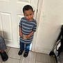 child, indoor, door, striped_shirt, blue_jeans, black_shoes, floor, tile, person, curious, standing, young_child, holding, expression, casual_clothing, home, wall, light_switch, shoe, footwear