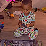 toddler, child, pajamas, toy, floor, play_mat, indoor, game_controller_prints, sitting, playing, wooden_floor, colorful_clothing, young_child, focus, casual, home, cute, small_toys, plastic_toy, checkered_pattern