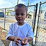 child, boy, white_tshirt, outdoor, chain_link_fence, sidewalk, daytime, sky, urban, casual_clothing, curious, short_hair, standing, hands, neighborhood, residential_area, young, person, street, fence