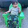 child, boy, green_chair, outdoor, concrete, vehicle, lollipop, casual_clothing, flip_flops, pants, gray_shirt, sitting, relaxed, hair, face, expression, footwear, daylight, plastic_chair, playful
