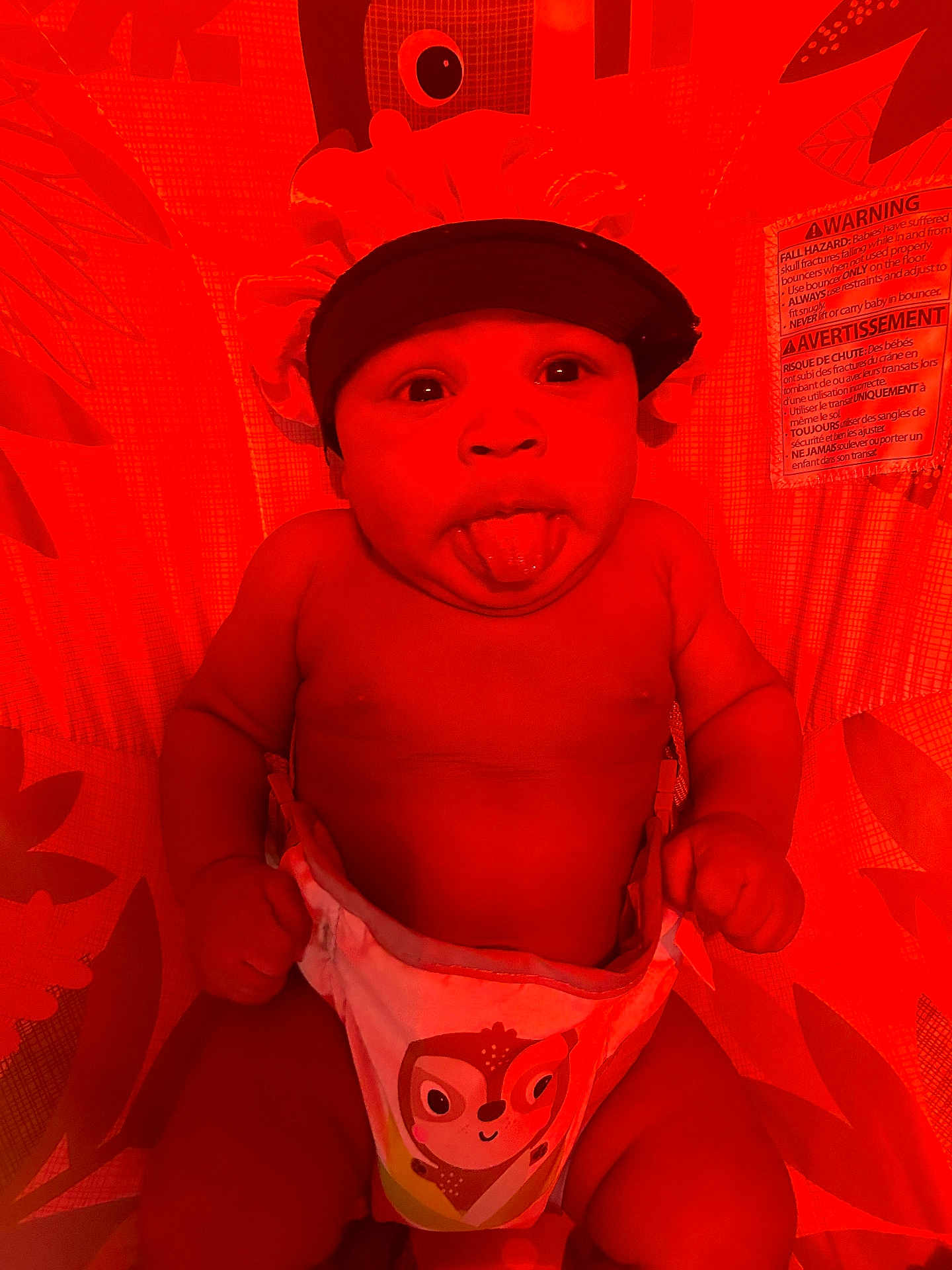 Johnathan is registered to the contest to win money with this photo: baby, infant, diaper, bonnet, tongue_out, playful, face, cute, child, patterned_background, owl_design, red_lighting, expression, skin, hand, lying_down, indoors, warning_label, fabric, closeup