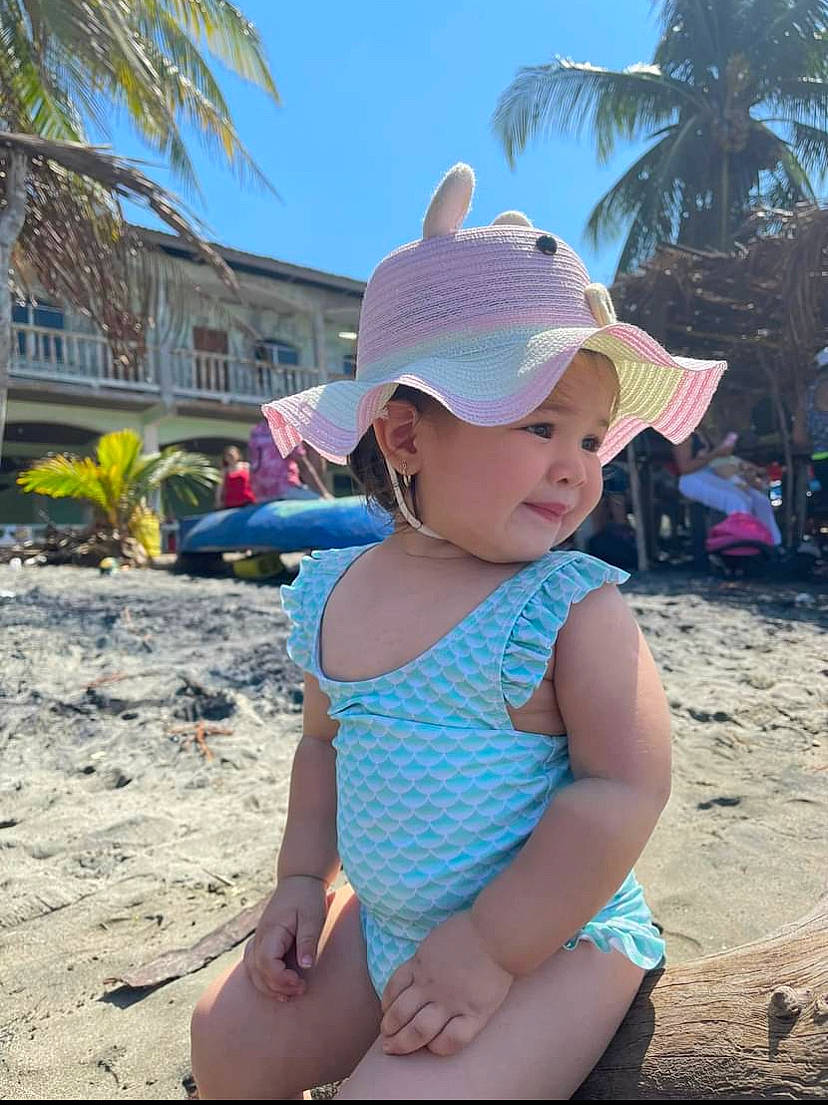 Annie is registered to the contest to win money with this photo: arecales, azure, baby, beach, body_of_water, electric_blue, event, fun, happy, hat, headgear, headwear, leisure, person, plant, sky, summer, sun_hat, swimwear, toddler