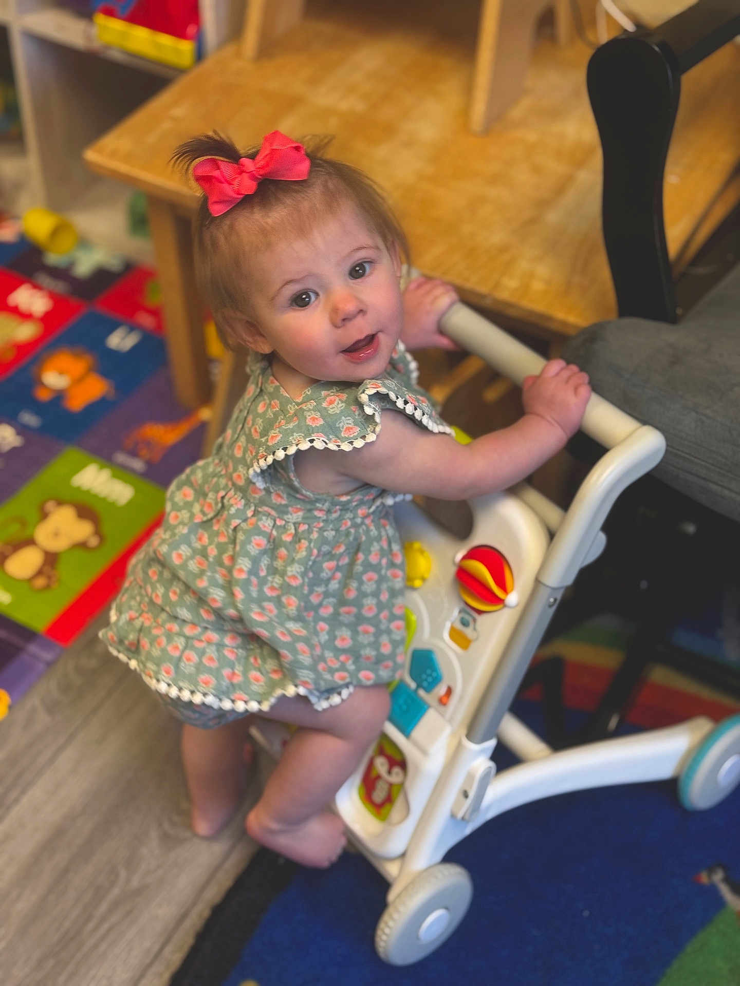 Alayna joined the competition — help win amazing prizes! toddler, child, baby_walker, pink_bow, green_dress, indoor, playroom, colorful_mat, wooden_furniture, curious_expression, barefoot, toy, flooring, person, smile, young_child, cute, playful, happy, early_learning