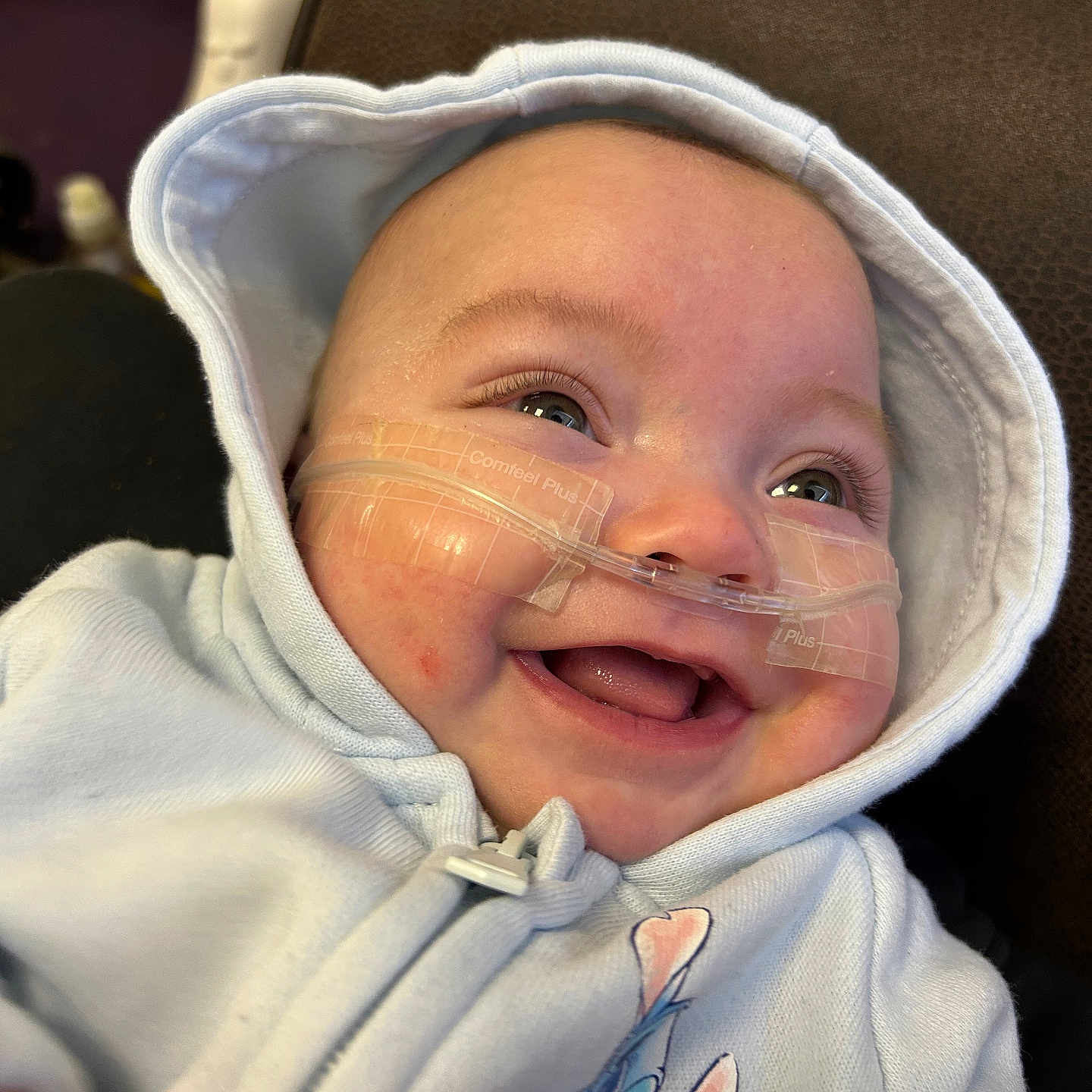 Hailey-Jo is registered to the contest to win money with this photo: baby, baby_clothes, blue_clothing, cartoon_character, child, close_up, cute, eyes, face, happy, hoodie, indoor, infant, medical_tape, mouth, nasal_cannula, person, skin, small_fingers, smiling