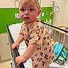Riley is registered to the contest to win money with this photo: baby, child, infant, hospital, hospital_bed, hospital_gown, cartoon_print, standing, face, portrait, wide_eyed, blond_hair, bed_rail, blanket, medical_room, indoor, colorful, cute, photography, safe