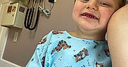 Riley joined the competition — help win amazing prizes! child, smiling, portrait, hospital, medical_exam_room, hospital_gown, medical_equipment, thermometer, wall, indoor, close_up, face, teeth, brown_hair, blue_clothing, cartoon_pattern, seated, camera, adult_arm, cheerful