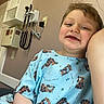 child, smiling, portrait, hospital, medical_exam_room, hospital_gown, medical_equipment, thermometer, wall, indoor, close_up, face, teeth, brown_hair, blue_clothing, cartoon_pattern, seated, camera, adult_arm, cheerful