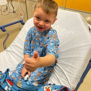 Riley joined the competition — help win amazing prizes! child, toddler, hospital, hospital_bed, pajamas, blanket, smiling, thumbs_up, portrait, indoor, medical_equipment, patient, bedroom, orange_wall, blue_clothing, animal_pattern, candid, seated, happy, healthcare