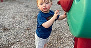 Riley joined the competition — help win amazing prizes! child, toddler, playground, play_equipment, slide, outdoor, park, portrait, standing, reaching, serious_expression, blue_shirt, gray_shorts, white_shoes, wood_chips, colorful, green_structure, red_structure, daylight, playtime