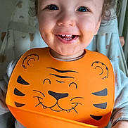 Ezio a rejoint le concours — aidez-le/la à gagner de superbes lots ! toddler, child, smile, blue_eyes, bib, orange_bib, tiger_design, messy_face, high_chair, indoor, happy, cute, baby, clothing, curly_hair, face, person, portrait, feeding_time, cozy