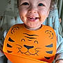 toddler, child, smile, blue_eyes, bib, orange_bib, tiger_design, messy_face, high_chair, indoor, happy, cute, baby, clothing, curly_hair, face, person, portrait, feeding_time, cozy