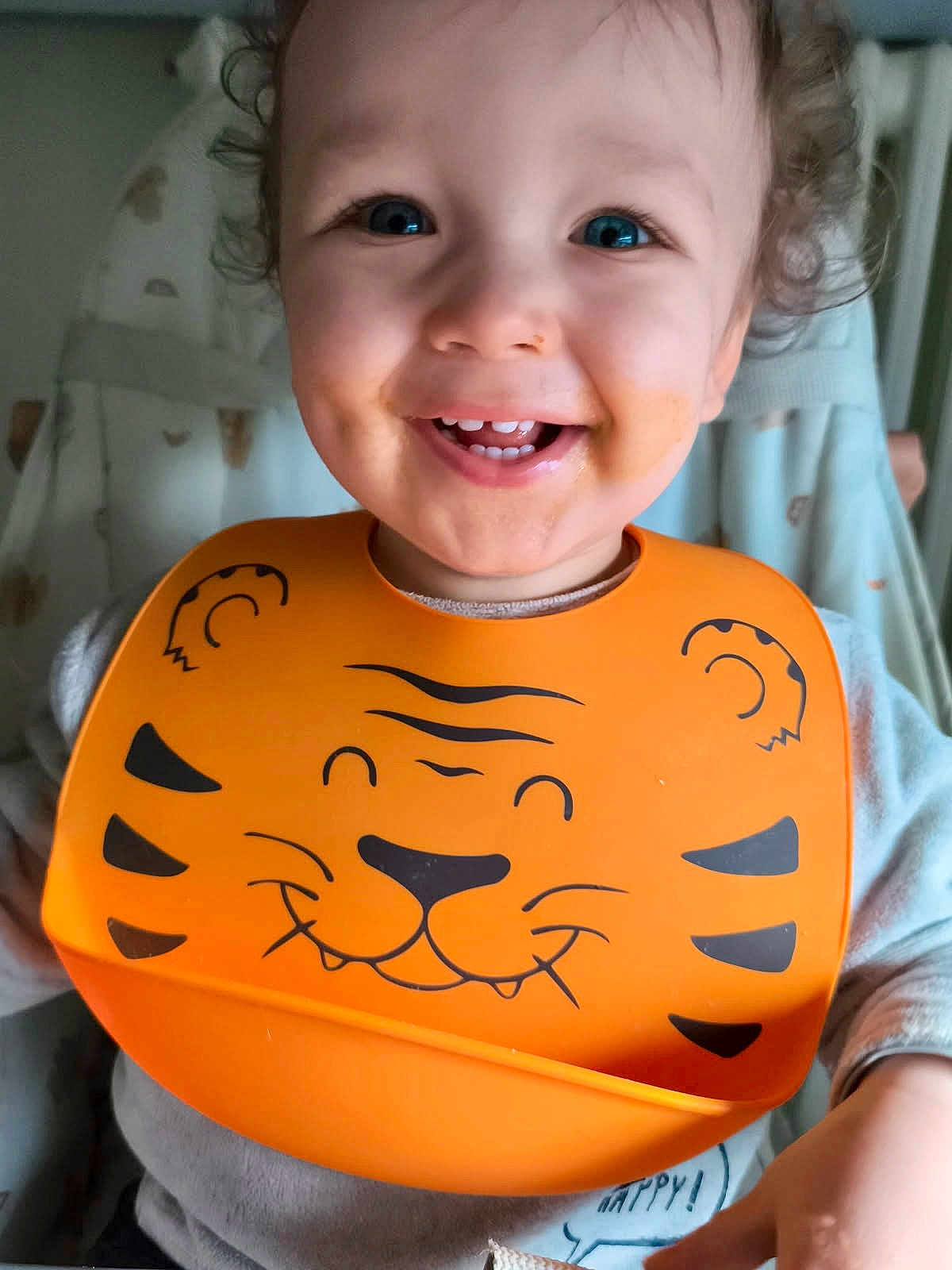 Ezio a rejoint le concours — aidez-le/la à gagner de superbes lots ! toddler, child, smile, blue_eyes, bib, orange_bib, tiger_design, messy_face, high_chair, indoor, happy, cute, baby, clothing, curly_hair, face, person, portrait, feeding_time, cozy