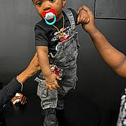 Jireh is registered to the contest to win money with this photo: baby, black_background, black_boots, casual_clothing, child, cute, denim, gold_chain, hands, holding, indoor, jewelry, overalls, pacifier, person, short_sleeve_shirt, standing, support, table, toddler