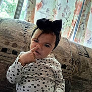Leilani is registered to the contest to win money with this photo: baby, infant, child, portrait, sitting, couch, bow, headband, sweater, patterned_clothing, leopard_print, nose_poking, hand, face, indoors, window, curtains, floral_curtains, natural_light, funny_pose
