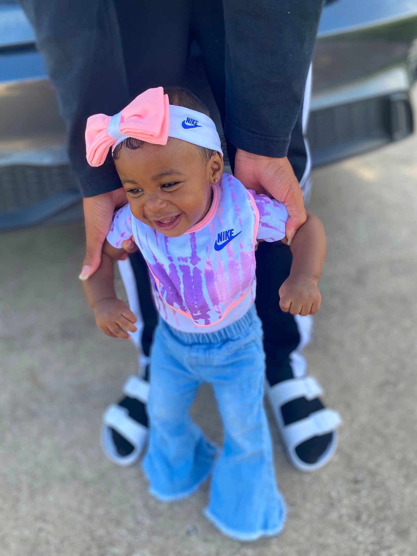 Ava is registered to the contest to win money with this photo: toddler, child, smiling, nike, headband, bow, adult_hands, jeans, outdoor, concrete, footwear, sandals, black_pants, colorful_clothing, baby, joyful, person, supporting, walking, fashion