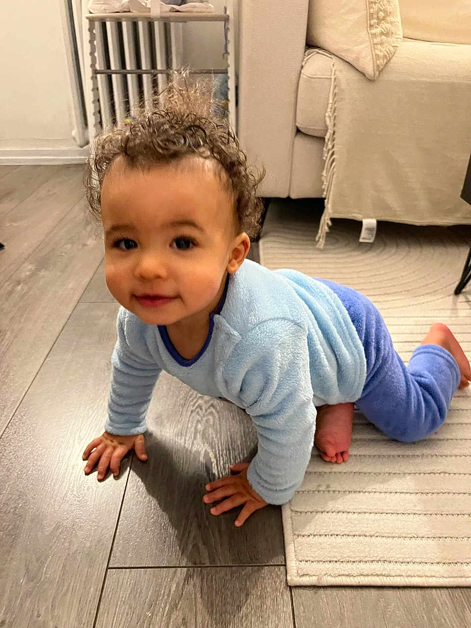 Isaac participe au concours pour gagner de l'argent avec cette photo : child, baby, toddler, crawling, curly_hair, blue_pajamas, hardwood_floor, area_rug, couch, living_room, smiling, hands, knees, bare_feet, radiator, furniture, indoor, portrait, playful, cute