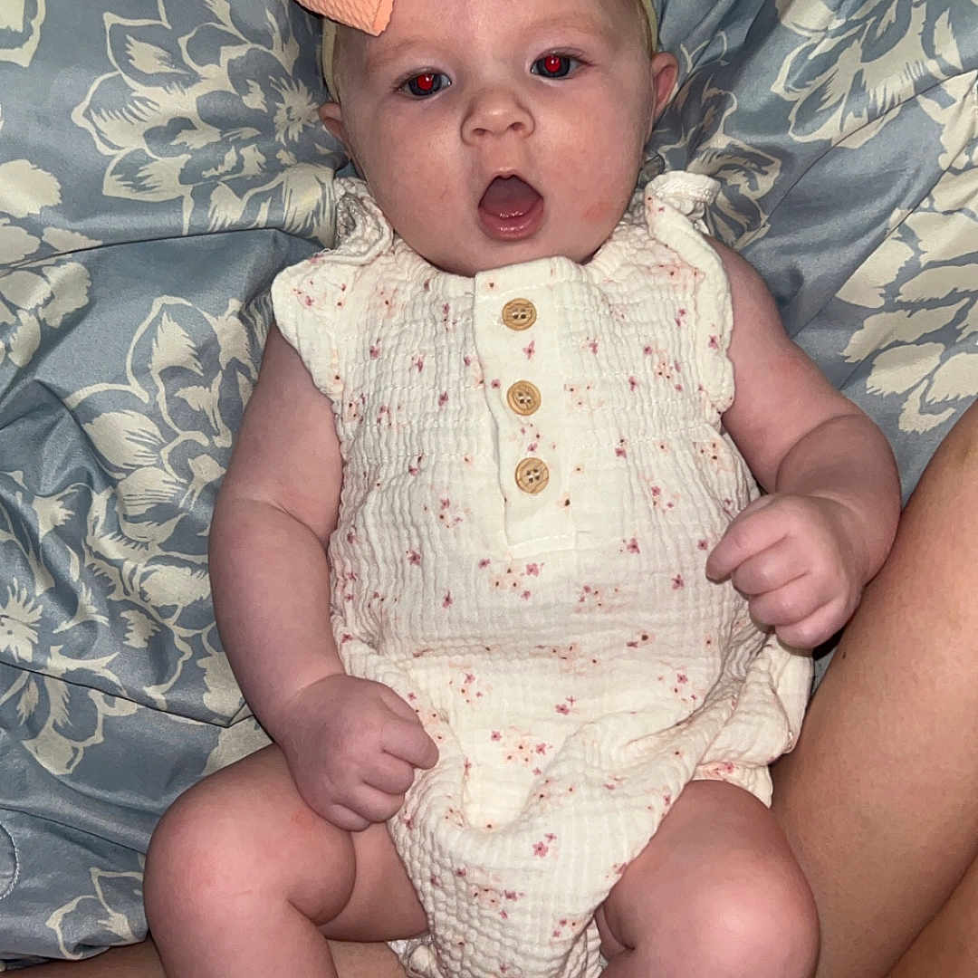 Josetta is registered to the contest to win money with this photo: arms, baby, bed, bow, child, clothing, comfort, fabric, floral, foot, hand, headband, infant, legs, onesie, pattern, person, sitting, skin, surprise