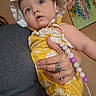 baby, child, hat, yellow_clothing, romper, pacifier, beads, hand, tattoo, ring, adult, indoor, person, portrait, blue_eyes, skin, holding, colorful_painting, floral_art, closeup