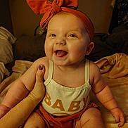Everleigh is registered to the contest to win money with this photo: baby, child, smiling, headband, bow, orange, clothing, hand, tattoo, skin, face, happy, sitting, indoors, blanket, person, infant, cute, warm_lighting, portrait