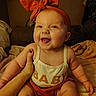 baby, child, smiling, headband, bow, orange, clothing, hand, tattoo, skin, face, happy, sitting, indoors, blanket, person, infant, cute, warm_lighting, portrait