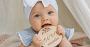 Everleigh is registered to the contest to win money with this photo: baby, infant, child, blue_eyes, white_bow, headband, blue_dress, lace, wooden_plaque, seven_months, chewing, cute, portrait, close_up, quilted_blanket, indoor, soft_light, smiling, hands, sitting