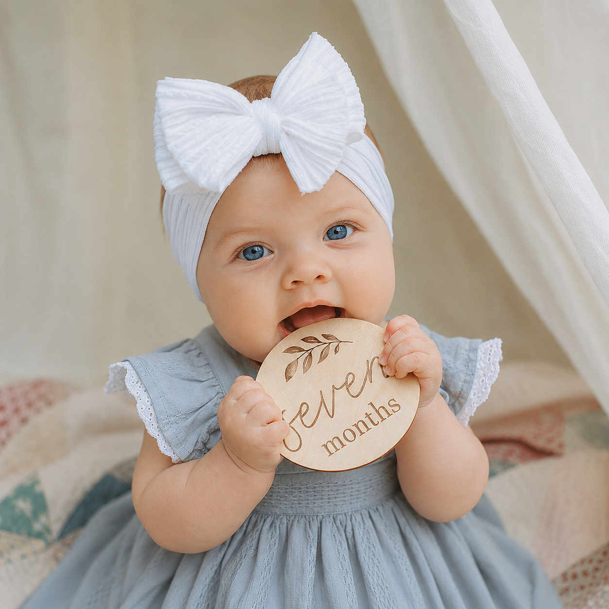 Everleigh is registered to the contest to win money with this photo: baby, infant, child, blue_eyes, white_bow, headband, blue_dress, lace, wooden_plaque, seven_months, chewing, cute, portrait, close_up, quilted_blanket, indoor, soft_light, smiling, hands, sitting
