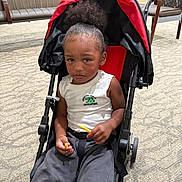 Ocean is registered to the contest to win money with this photo: child, toddler, stroller, portrait, sitting, serious_expression, curly_hair, ponytail, red_canopy, white_shirt, gray_pants, snack, yellow_food, seatbelt, indoor, waiting_room, chairs, carpet, close_up, photography