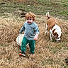 toddler, child, dog, grass, field, bucket, outdoor, pet, walking, curly_hair, green_pants, grey_shirt, sneakers, animal, nature, play, young_child, canine, grassland, casual_clothing