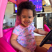 Kailani is registered to the contest to win money with this photo: child, toddler, smiling, toy_car, pink, curly_hair, indoor, steering_wheel, playful, happy, clothing, purple_shirt, unicorn_pattern, furniture, television, stacked_items, wooden_floor, person, cute, portrait