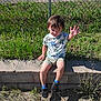 Noah Carter joined the competition — help win amazing prizes! child, dinosaur_shirt, shorts, sitting, waving, grass, chain_link_fence, brick_wall, outdoor, sunlight, shadow, playful, casual_clothing, footwear, blue_socks, person, nature, daylight, smiling, young