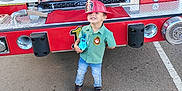 Noah Carter is registered to the contest to win money with this photo: child, firefighter, fire_truck, toy, red_hat, jeans, sneakers, smiling, outdoor, parking_lot, vehicle, uniform, badge, asphalt, front_view, daylight, playing, cute, boy, standing