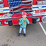 child, firefighter, fire_truck, toy, red_hat, jeans, sneakers, smiling, outdoor, parking_lot, vehicle, uniform, badge, asphalt, front_view, daylight, playing, cute, boy, standing