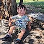 child, boy, dinosaur_shirt, shorts, black_shoes, tree, shade, concrete, cracks, grass, outdoor, sunlight, smiling, casual_clothing, park, nature, playful, young_child, happy, summer
