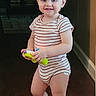 toddler, child, barefoot, striped_onesie, toy, indoor, wooden_floor, hallway, smile, blond_hair, person, young_child, standing, playful, casual_clothing, home, cute, happy, innocent, portrait