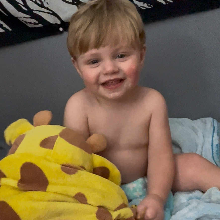 Dryker joined the competition — help win amazing prizes! toddler, child, smiling, bed, blanket, plush_toy, giraffe, yellow, brown_spots, indoor, portrait, happy, casual, hair, face, diaper, gray_wall, artwork, soft_texture, cute