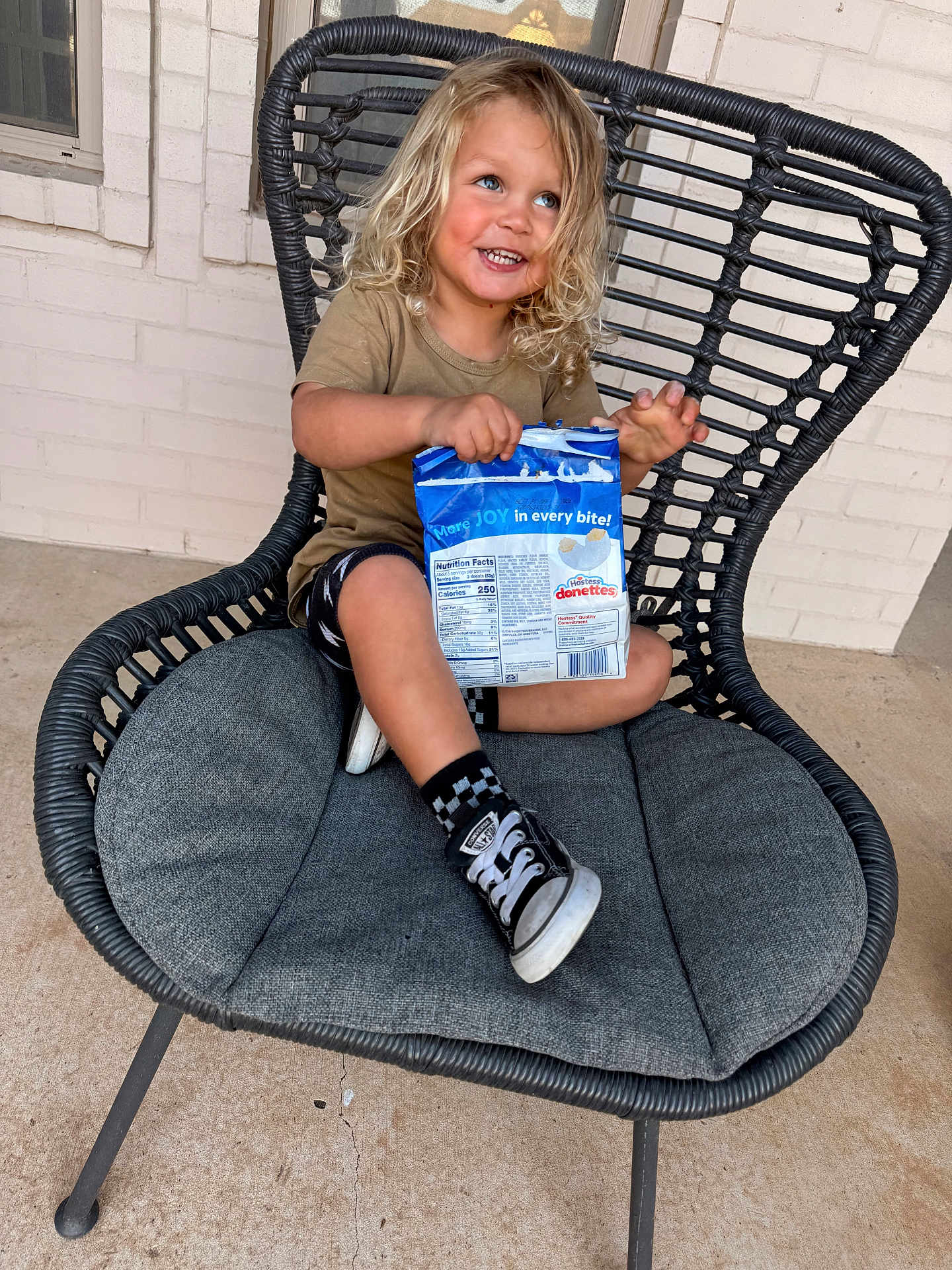 Kameron is registered to the contest to win money with this photo: child, toddler, smiling, curly_hair, blonde_hair, snack, snack_package, chair, wicker_chair, cushion, sneaker, checkered_socks, porch, brick_wall, window, outdoor, sitting, messy_hair, candid_photo, joyful