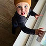 child, toddler, baby, beanie, blue_eyes, sticking_tongue, onesie, long_sleeve, hand, window, windowsill, wood_floor, indoor, portrait, looking_up, standing, cute, playful, hat, curious