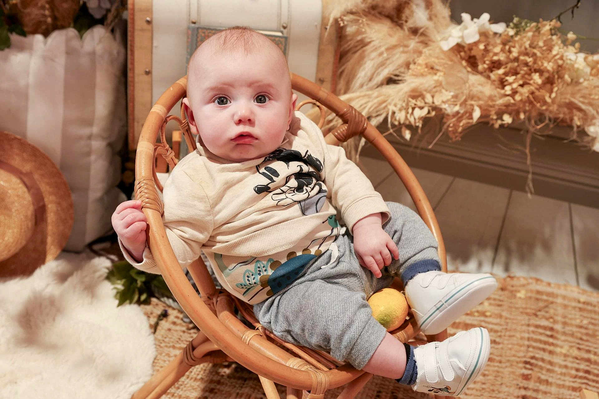 Lucas a rejoint le concours — aidez-le/la à gagner de superbes lots ! baby, infant, child, rattan_chair, plush_rug, straw_hat, dried_flowers, lemon, sneakers, socks, sweatpants, long_sleeve_shirt, wide_eyes, curious_expression, indoor, home_decor, wooden_floor, plant, portrait, seated