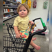 Ethan is registered to the contest to win money with this photo: toddler, child, shopping_cart, store, clothing, yellow_shirt, winnie_the_pooh, shorts, barefoot, curly_hair, blue_eyes, floor, aisle, hand, face, person, retail, indoor, grocery, cute