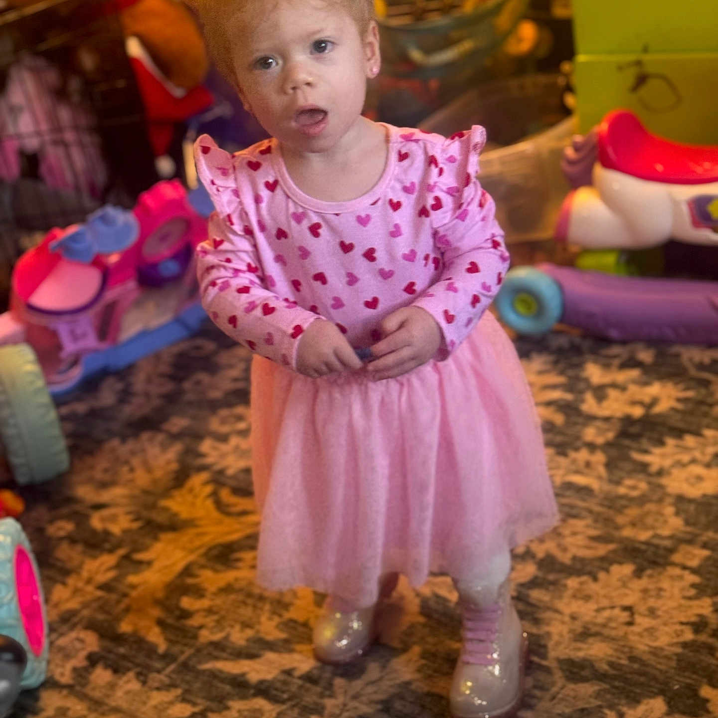 Magnolia-Diane is registered to the contest to win money with this photo: boots, carpet, child, clothing, curly_hair, expression, floor, heart_pattern, indoor, indoor_lighting, patterned_rug, person, pink_dress, plastic_toy, playroom, standing, storage_unit, toddler, toys, young_child