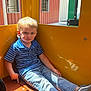 child, boy, blonde_hair, blue_shirt, striped_shirt, jeans, sneakers, yellow_wall, window, building, red_wall, white_wall, sitting, indoor, sunlight, shadow, contemplative, casual_clothing, young, portrait