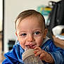 child, baby, infant, toddler, bottle, feeding_bottle, feeding, smile, happy, blue_clothing, highchair, face, eyes, hand, mouth, indoor, portrait, close_up, blurred_background, hair