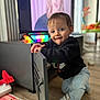 child, toddler, smile, indoor, living_room, television, toy, hardwood_floor, sneakers, jeans, sweatshirt, cabinet, television_screen, colorful_light, playmat, deer_toy, curtains, furniture, portrait, bokeh