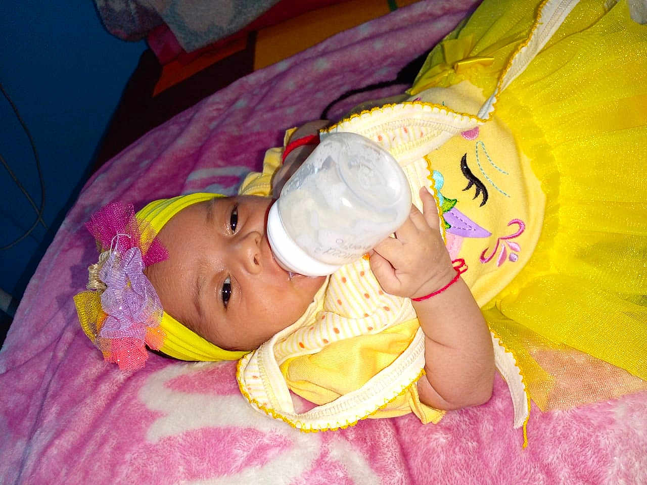 Crystal is registered to the contest to win money with this photo: baby, bottle, milk, yellow_clothing, headband, flower, blanket, infant, child, cute, lying_down, feeding, hand, pink_blanket, bed, indoors, person, sleepwear, toddler, smiling
