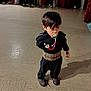 child, pacifier, black_clothing, patterned_belt, floor, people, costumes, group, indoor, shoes, standing, traditional_clothing, crowd, blurred_background, person, young_child, hair, face, shadow, event