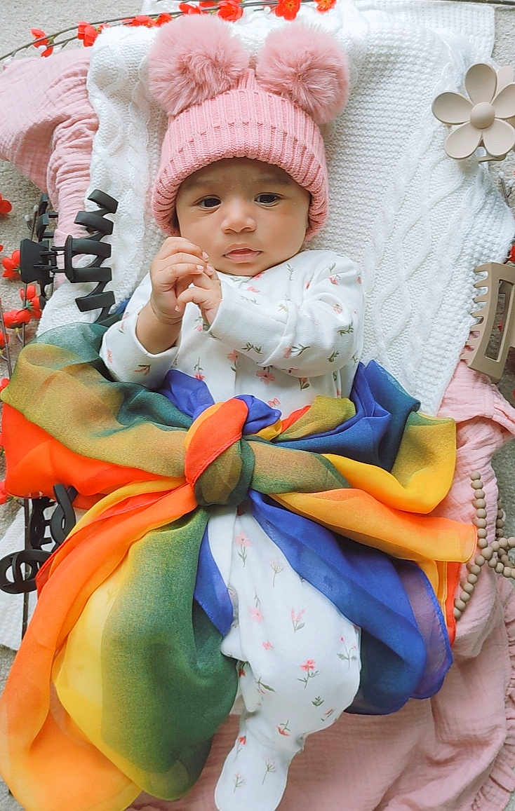 Nayra is registered to the contest to win money with this photo: baby, infant, pink_hat, pom_poms, floral_onesie, colorful_scarf, blanket, soft_toys, cute, child, indoors, portrait, smiling, cozy, warm, decorations, playful, lying_down, headwear, newborn