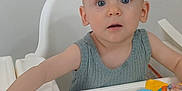 Noah is registered to the contest to win money with this photo: baby, child, high_chair, toy, blue_eyes, curious, indoor, white, gray_clothing, sleeveless, table, face, person, young_child, floor, furniture, arm, hand, foot, expression