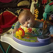 Slade is registered to the contest to win money with this photo: activity_walker, baby, blanket, child, clothing, colorful, cushion, cute, face, furniture, head, home, indoor, infant, pacifier, person, plaything, seated, sleepy, toy
